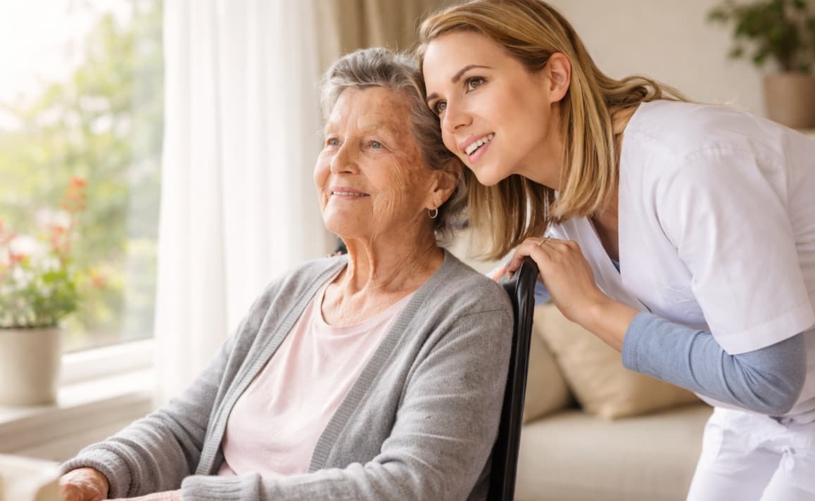 A caregiver and an elderly woman smile amidst colorful blooming flowers in a garden, conveying warmth and companionship. Bright, uplifting atmosphere.